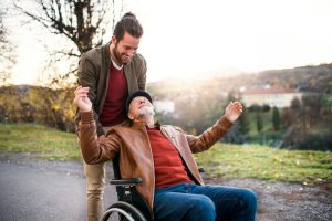 Young Man and Senior Father In Wheelchair Walking During Sunset